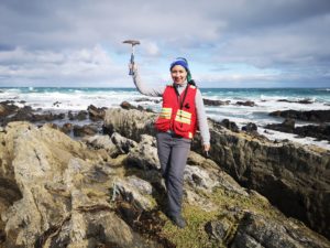 Karla standing on a rocky beach holding a geologists pick
